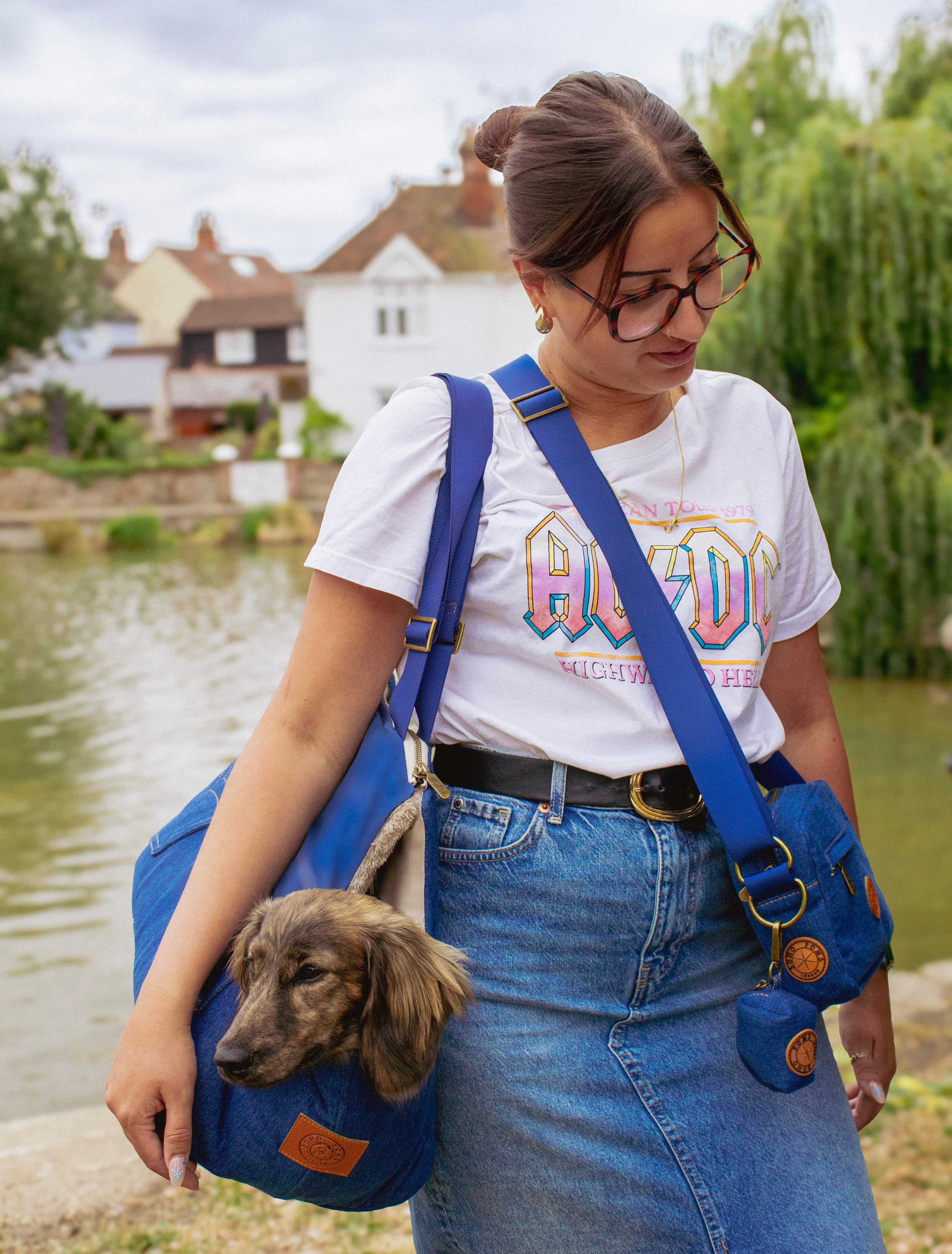 A woman wearing a white AC/DC band t-shirt and denim skirt carries a small dachshund dog in a blue pet shoulder bag with a matching crossbody bag, set against a background of a river and houses.